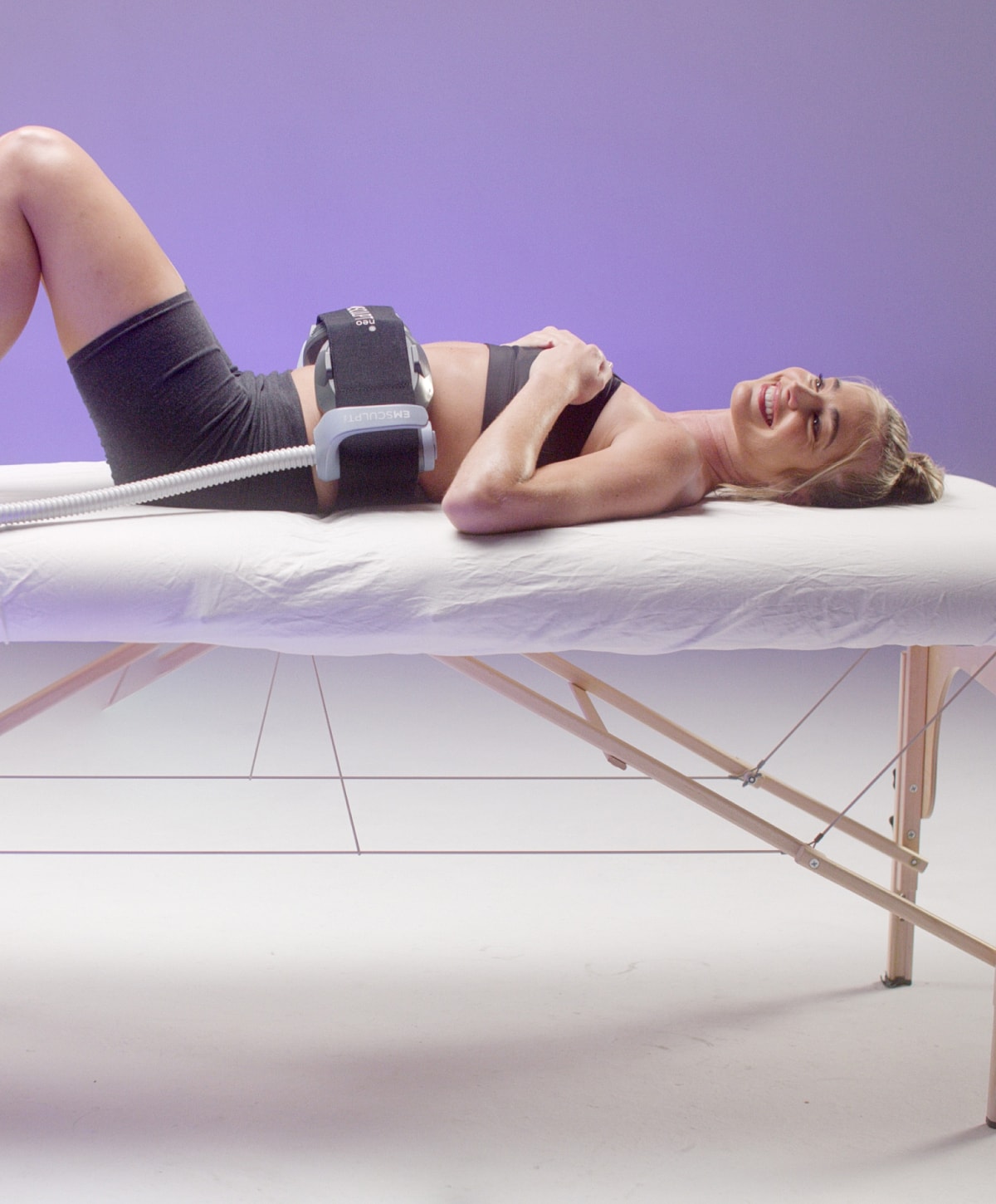 emsculpt new patient model laying on a cot smiling with the emsculpt neo device on her abdomen