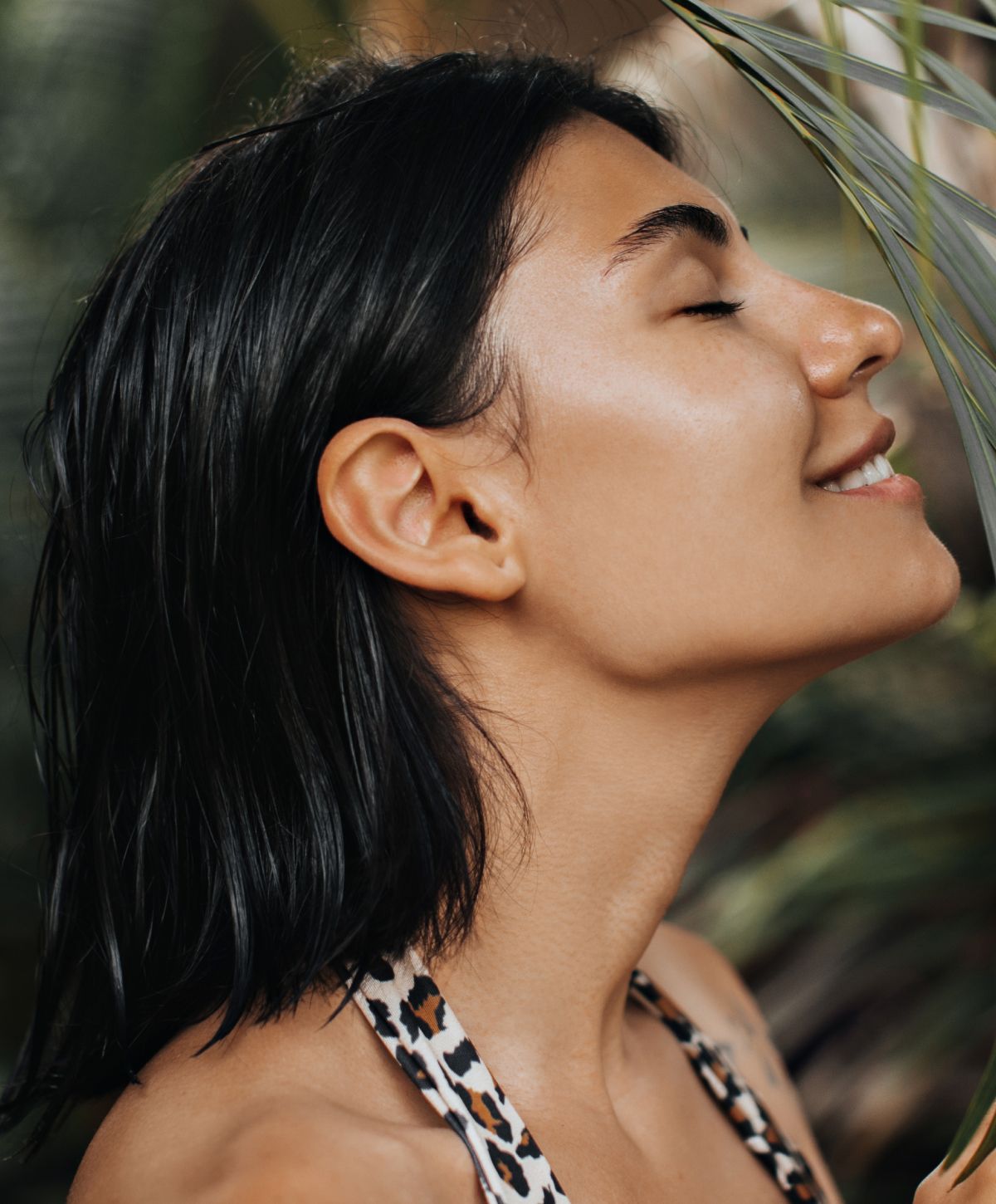 Profile of an accutite patient model smiling with her eyes closed amidst green foliage.